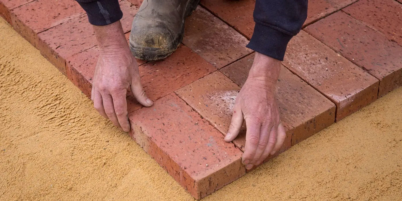 Hands laying red paver bricks on a sand base