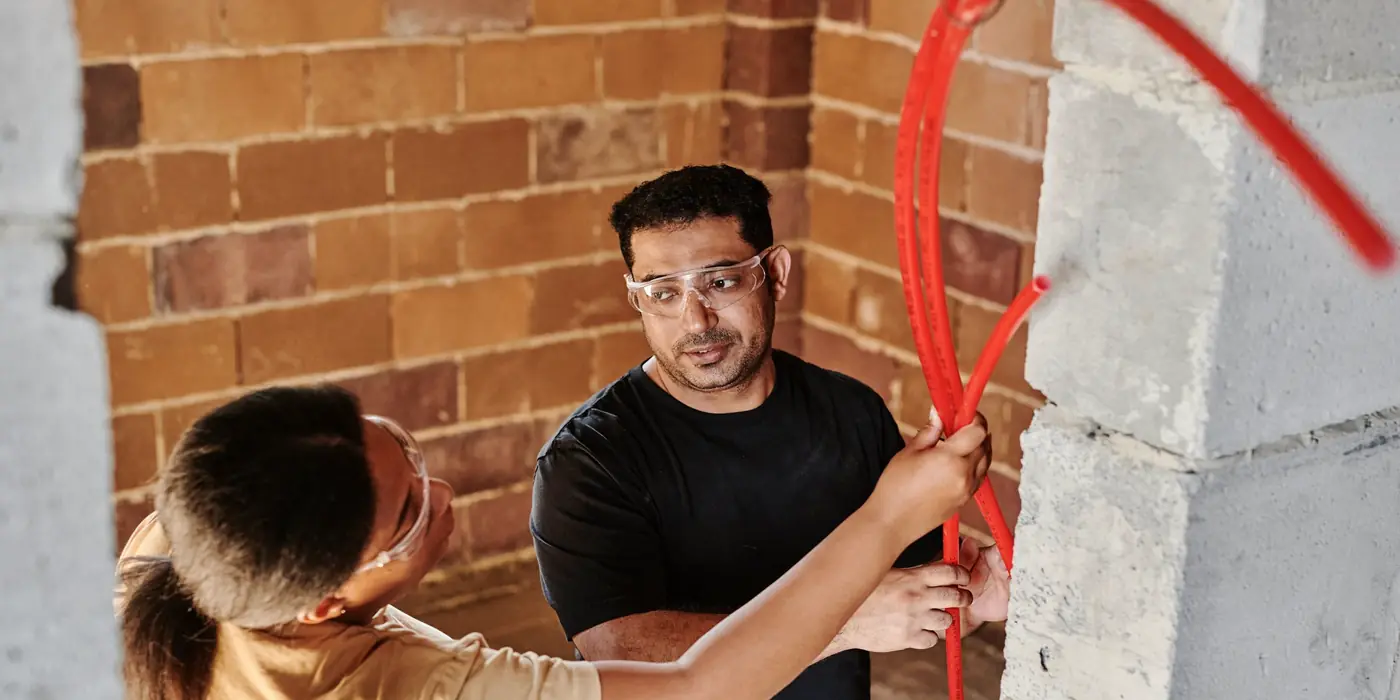 Two workers fitting red water supply tubing along a brick wall