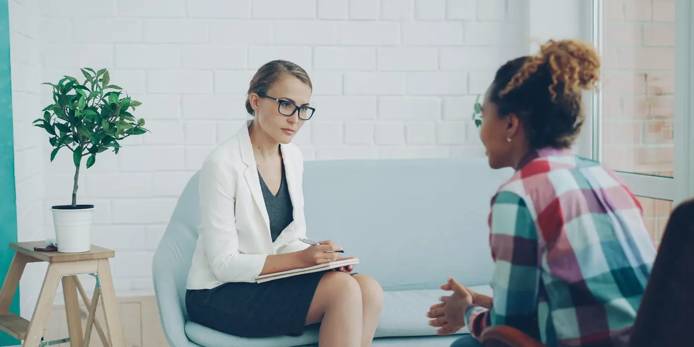 Clinician with a notebook listening to a patient in a bright office with a plant and a light blue sofa